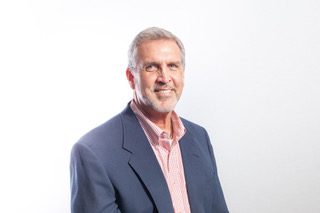 Middle-aged man with gray hair and beard wearing blue blazer and pink checkered shirt on white background
